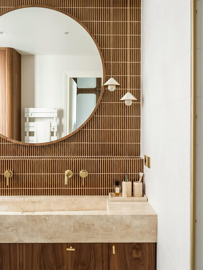 Meuble vasque en pierre, miroir rond et carrelage vertical brun avec robinetterie laiton — rénovation salle de bain ADJ Intérieurs